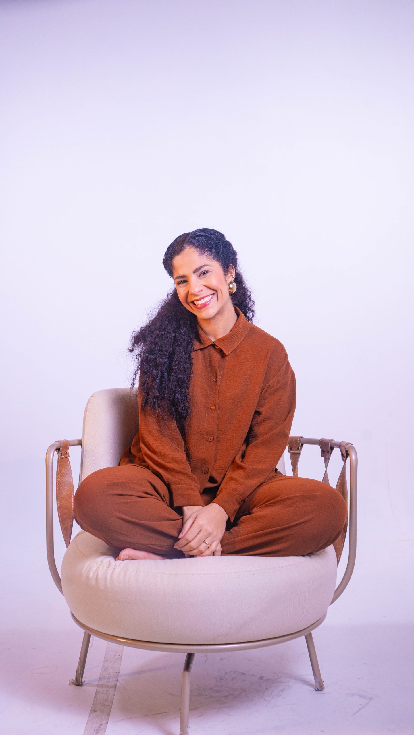 Dra. Sara Dutra sentada de pernas cruzadas numa poltrona, com look terracota