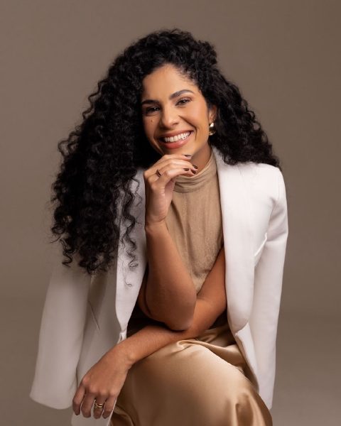 Dra. Sara Dutra sentada num banquinho, de blazer branco e saia champanhe, sorrindo com a mão no queixo